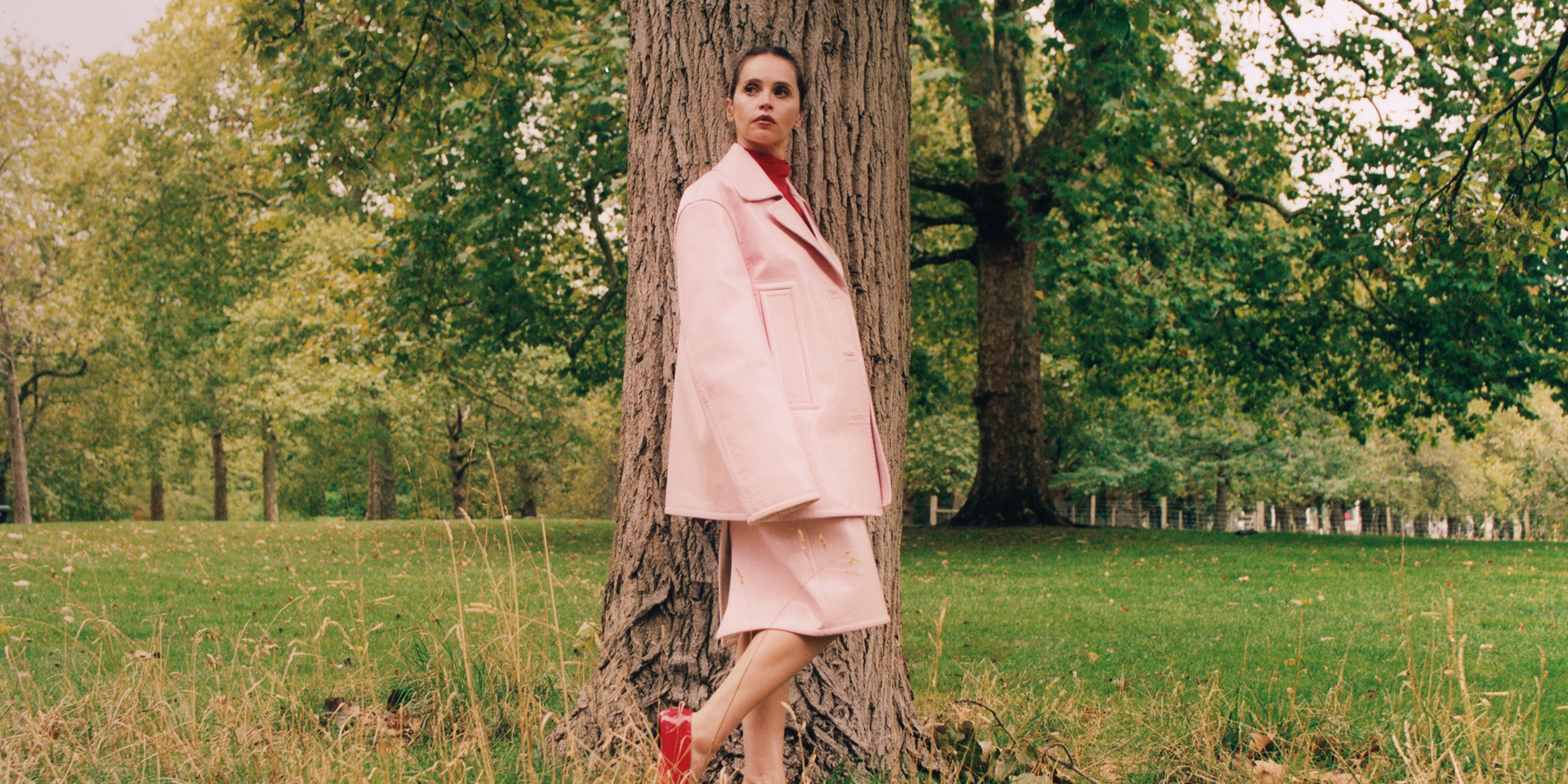 The actor Felicity Jones in a pink coat in a park.