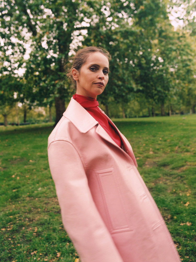 The actor Felicity Jones in a pink coat in a park.