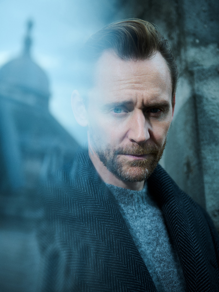 Tom Hiddleston looking at the camera with a building reflected in a window behind him.